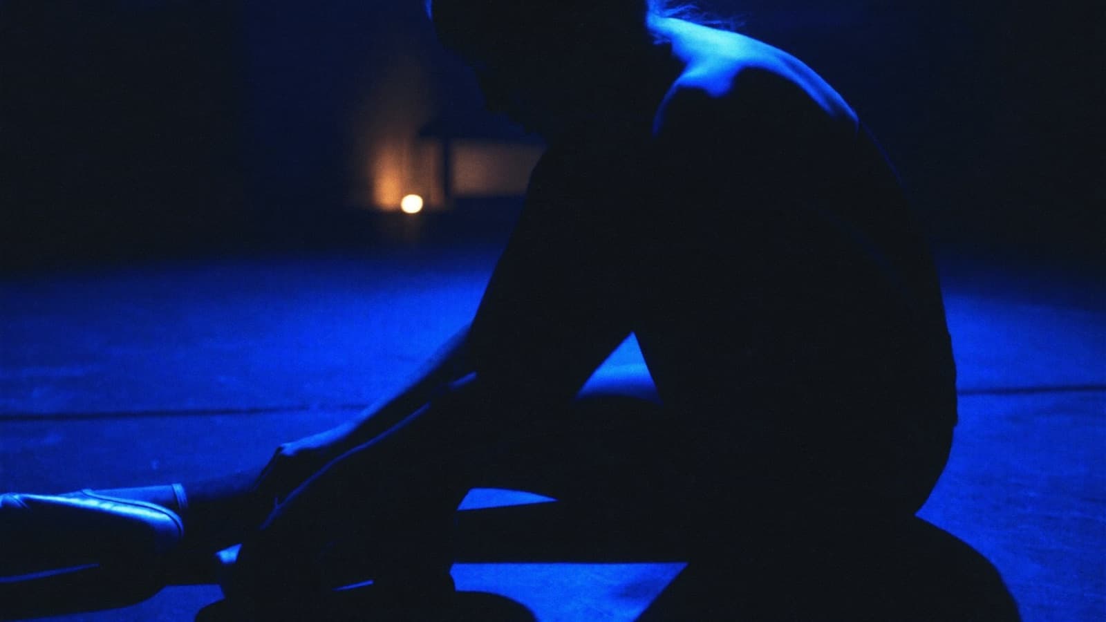 A dancer tying pointe shoes on a dark studio floor, lit by a cobalt rim light